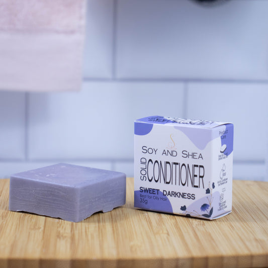 On a wooden tray sits a purple bar of conditioner next to a white box with a purple abstract pattern reading SOLID CONDITIONER against a blurred tiled background.