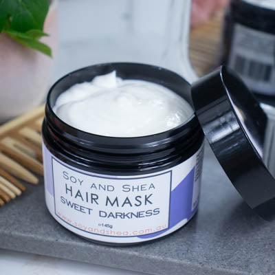 An overhead shot of a black jar with the lid resting on its side revealing a white cream inside, sits on a dark grey tile surface against a blurred background of a bathroom counter top. The jar features a white label with purple abstract design with text HAIR MASK SWEET DARKNESS.