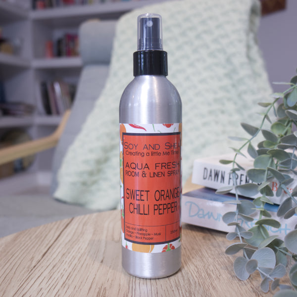 A metal bottle with spray nozzle and orange label sits on a wooden surface next to a stack of books and greenery. Behind is a chair with blanket and a blurred book case.