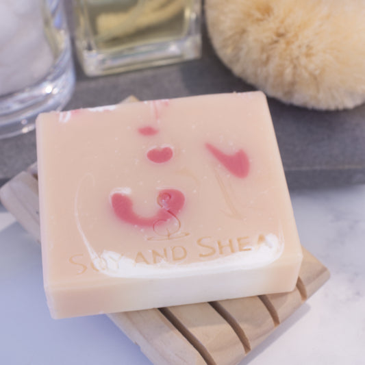 A pale orange soap bar with red and white drops of colour lays flat on a wooden soap dish next to a stone surface with bottles.