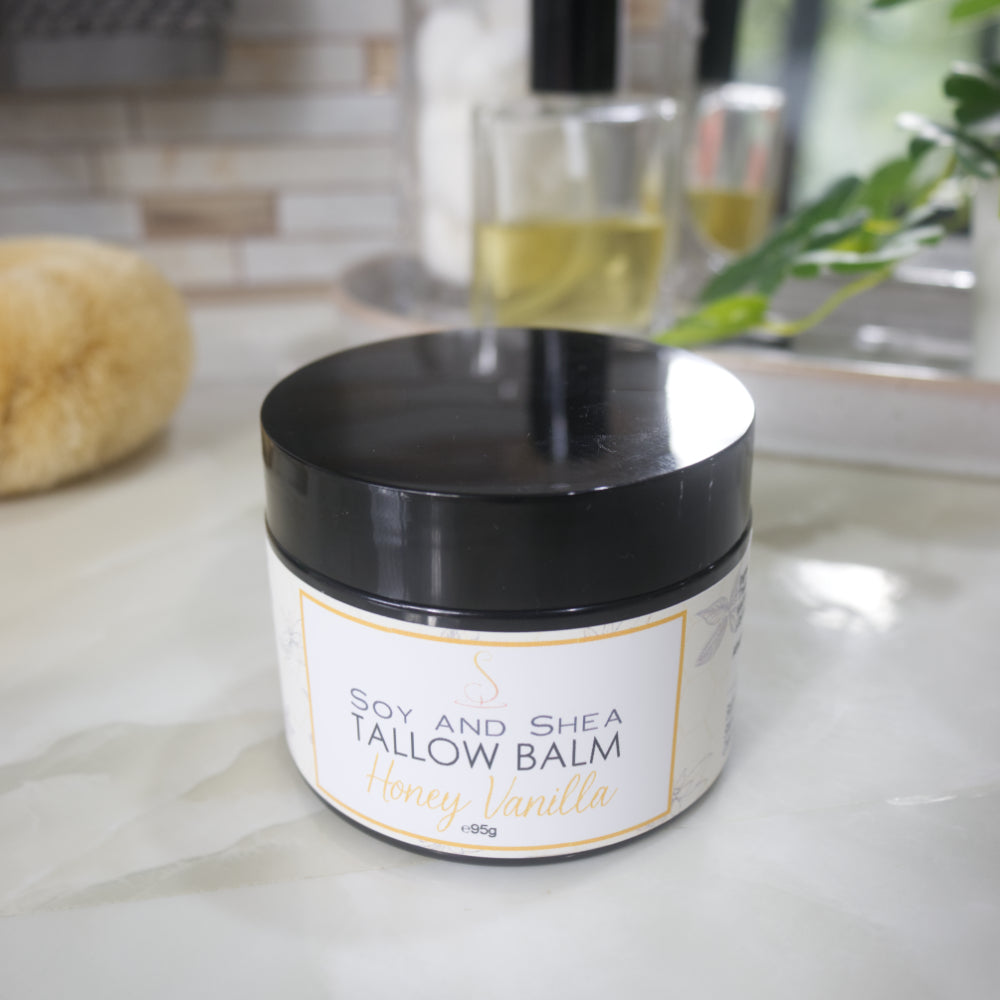 A black jar with a decorative label is displayed on a bathroom countertop. The label identifies it as Tallow Balm in Honey Vanilla Scent.