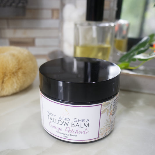 A black jar with a decorative label is displayed on a bathroom countertop. The label identifies it as Tallow Balm in Orange and Patchouli.
