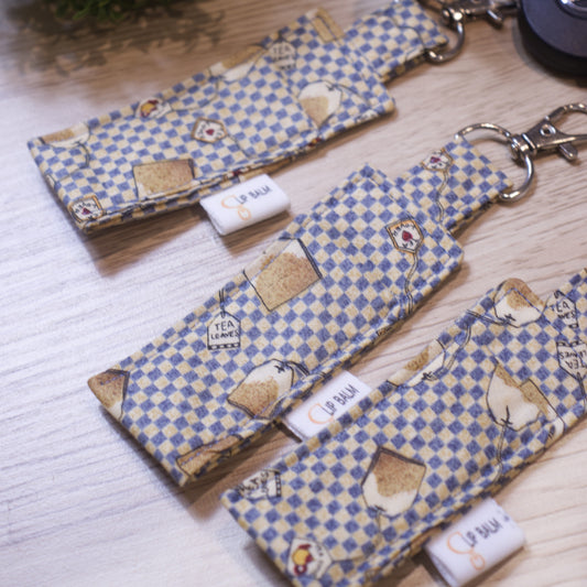 A selection of three lip balms showing variations in fabric design, lie on a wooden surface. The Clip balms have a blue and cream small checkered pattern with randomly placed teabags