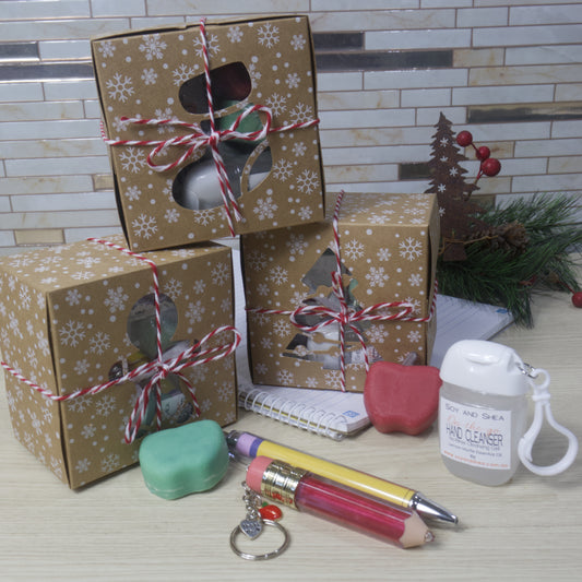 Three kraft boxes with white snowflakes and tied with red twine are stacked on a wood bench showing various window cut outs in christmas themes. In front sits two apple shaped soaps in red and greem a pencil shaped lip oil on key chain, a decorative pen and a bottle of hand cleanser