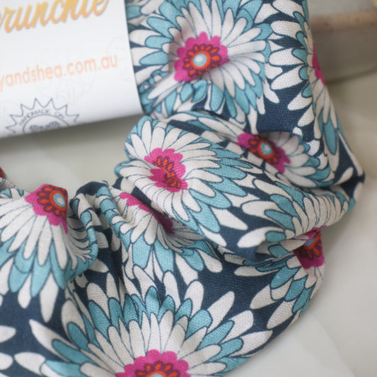 A close up image of the Teal Gerbera scrunchie showing pattern detail. The scrunchie gerbera like flowers in shades of dark blue, white and teal with bright pink centers.