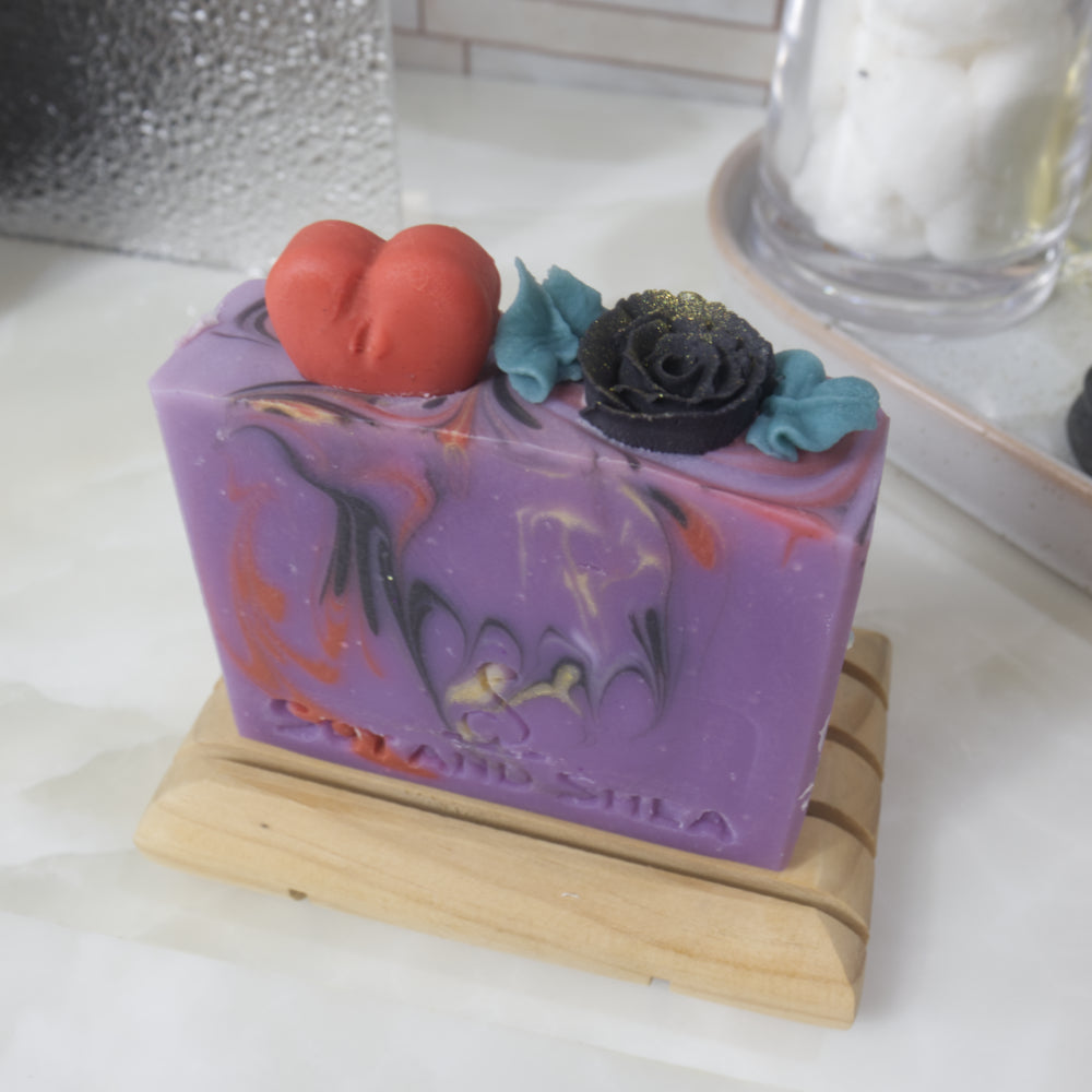 A soap bar stands on a wooden soap rack on a bathroom counter surrouned by perfume, a jar with cotton balls and a plant. The soap is bright purple with swirls of red, gold and back. The image focuses on the top of the soap which has embellishments made of soap in the shape of a red heart and a black rose with green leaves.