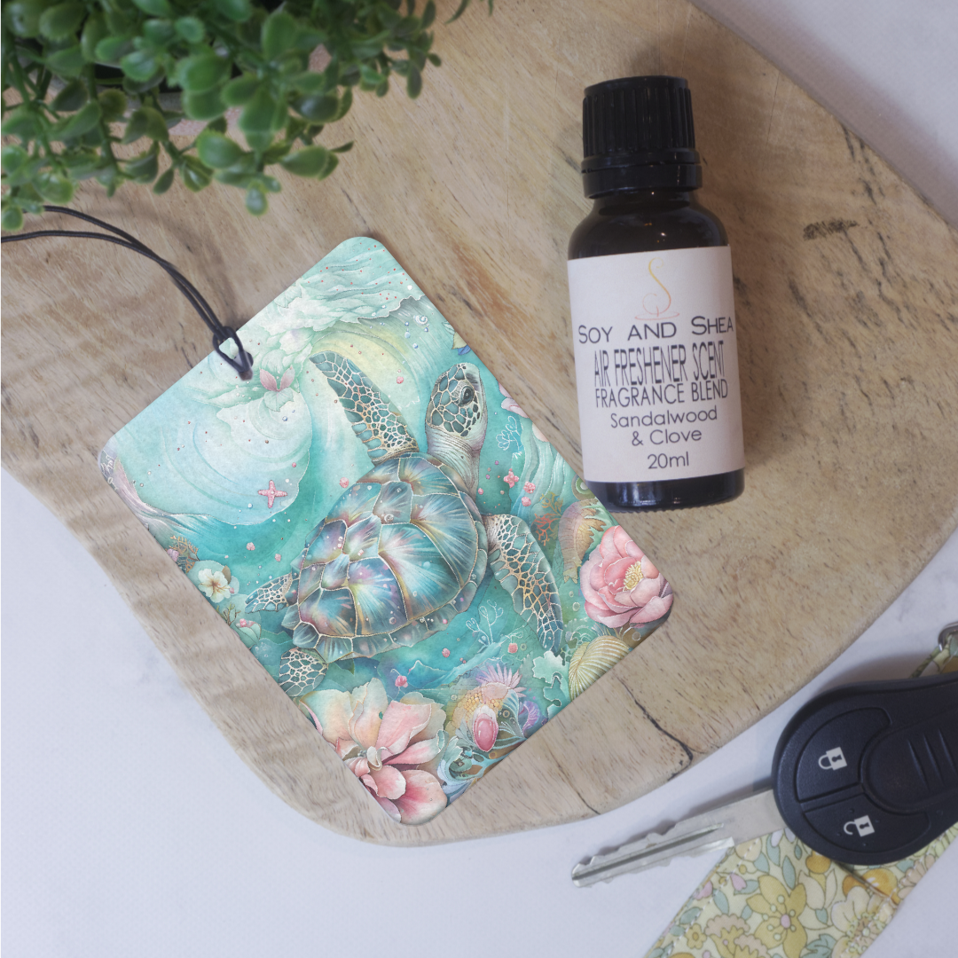 A car air freshener featuring an artistic depiction of a turtle surrounded by floral elements, placed on a wooden surface with various items in the background. The design has a boho feel in colours of soft pink and teal