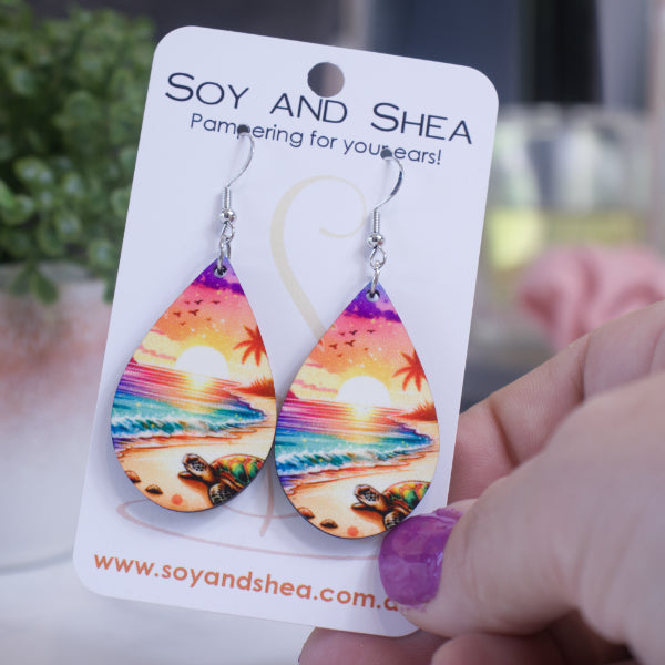 A hand holds a white card branded with SOY AND SHEA with a pair of earrings hanging on hooks. The earrings are teardrop shaped depicting a beach at sunset. In the foreground, a turtle can be seen crawling across the sand towards the water which has shades of teal to pink-purple. A sun is seen setting on the horizon with a sky in ombre colour from orange to pink then purple