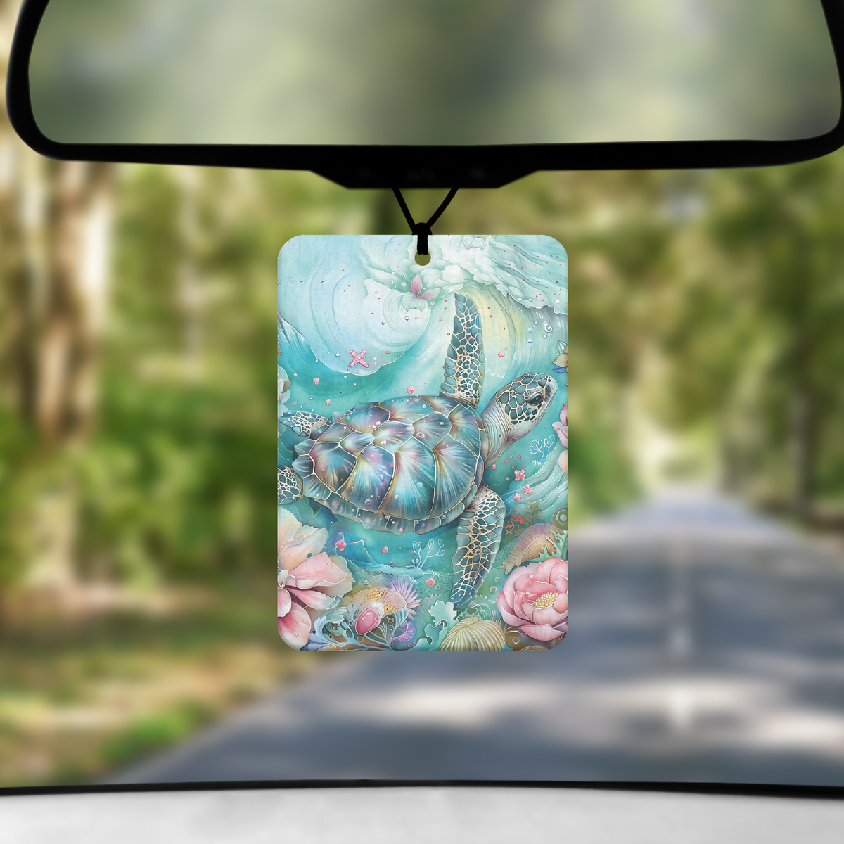 A mock up image showing a view through a car windscreen with a long tree lined road. From the rearview mirror hangs an air freshener with a boho style image of a turtle swimming through a reef of flowers.
