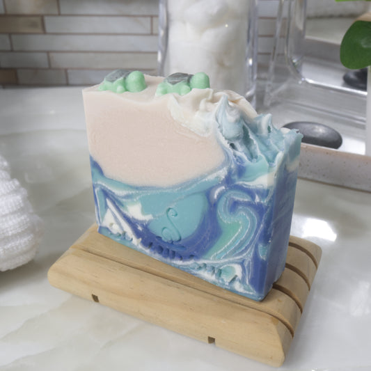 A bar of soap stands on a wooden soap rack on a bathroom counter top. The soap features a swirling pattern of ocean blues with a beige left corner. On top of the beige section is two small turtle shapes