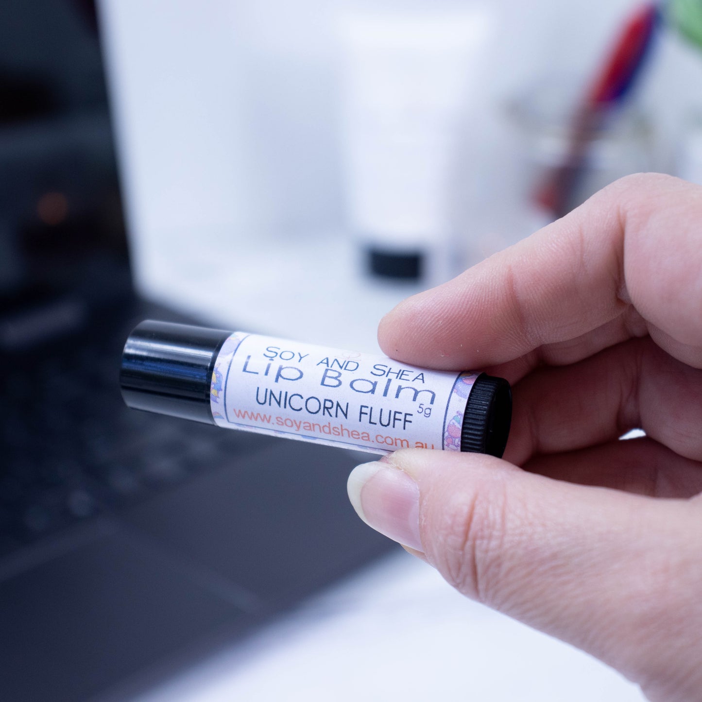 A hand holds a black tube of lip balm against a blurred background with an open laptop computer on a white surface. The label has text LIP BALM UNICORN FLUFF against a white background with a border of pale colours