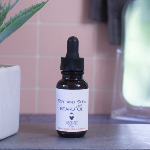 A brown bottle with white label and black dropper lid stands on stone surface next to a pot with green leaves against a blurred tiled background. The label reads BEARD AND MO OIL and shows the image of a man with bead and moustache.