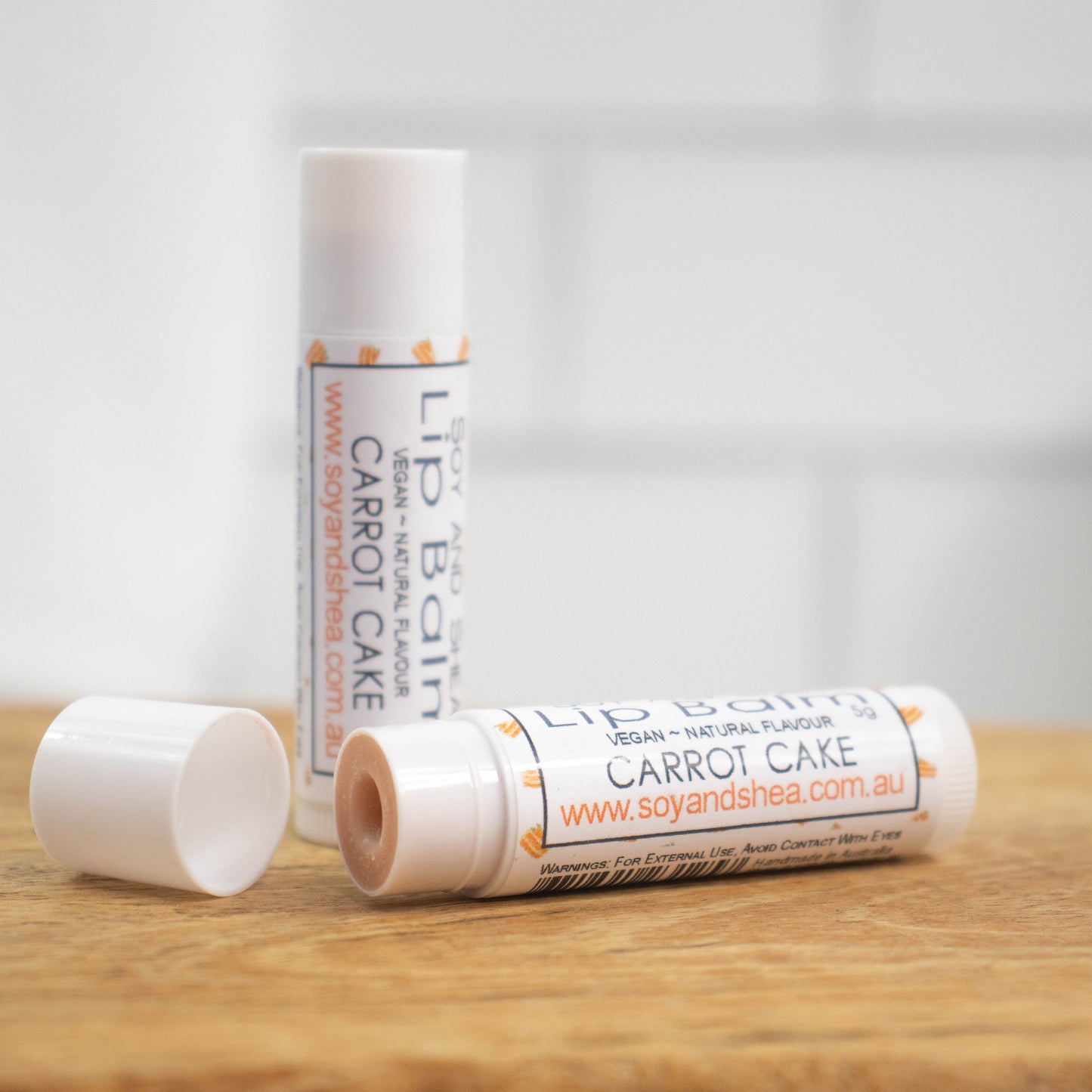 Two white tubes of lip balm are displayed on a wooden surface with blurred tiled background. One tube stands on its end while the other lays flat in front with the cap removed revealing a tan balm. The label is white with illustrations of pieces of cake