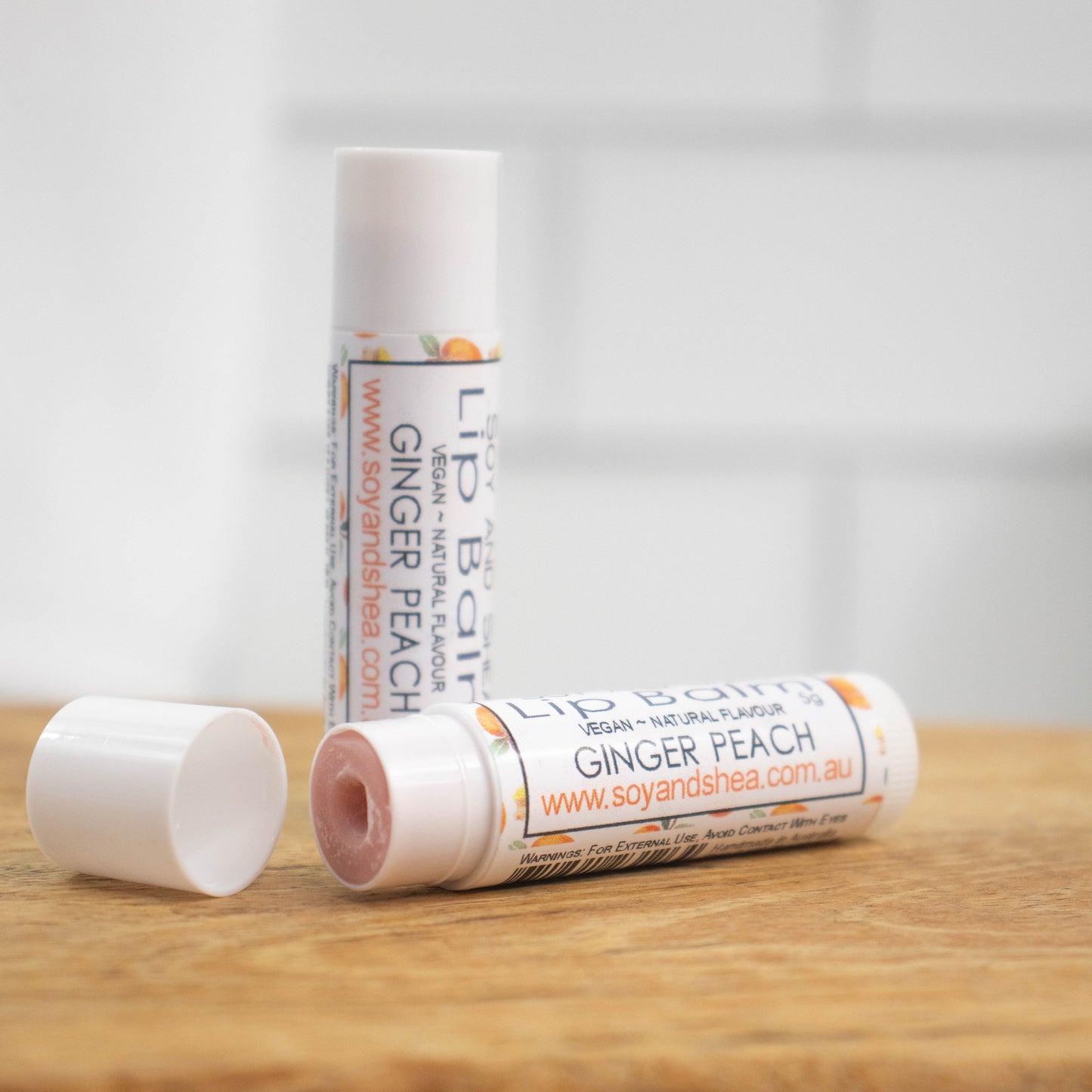 Two white tubes of lip balm are displayed on a wooden surface with blurred tiled background. One tube stands on its end while the other lays flat in front with the cap removed revealing a light brown balm. The label is white with peach and ginger pieces randomly scattered
