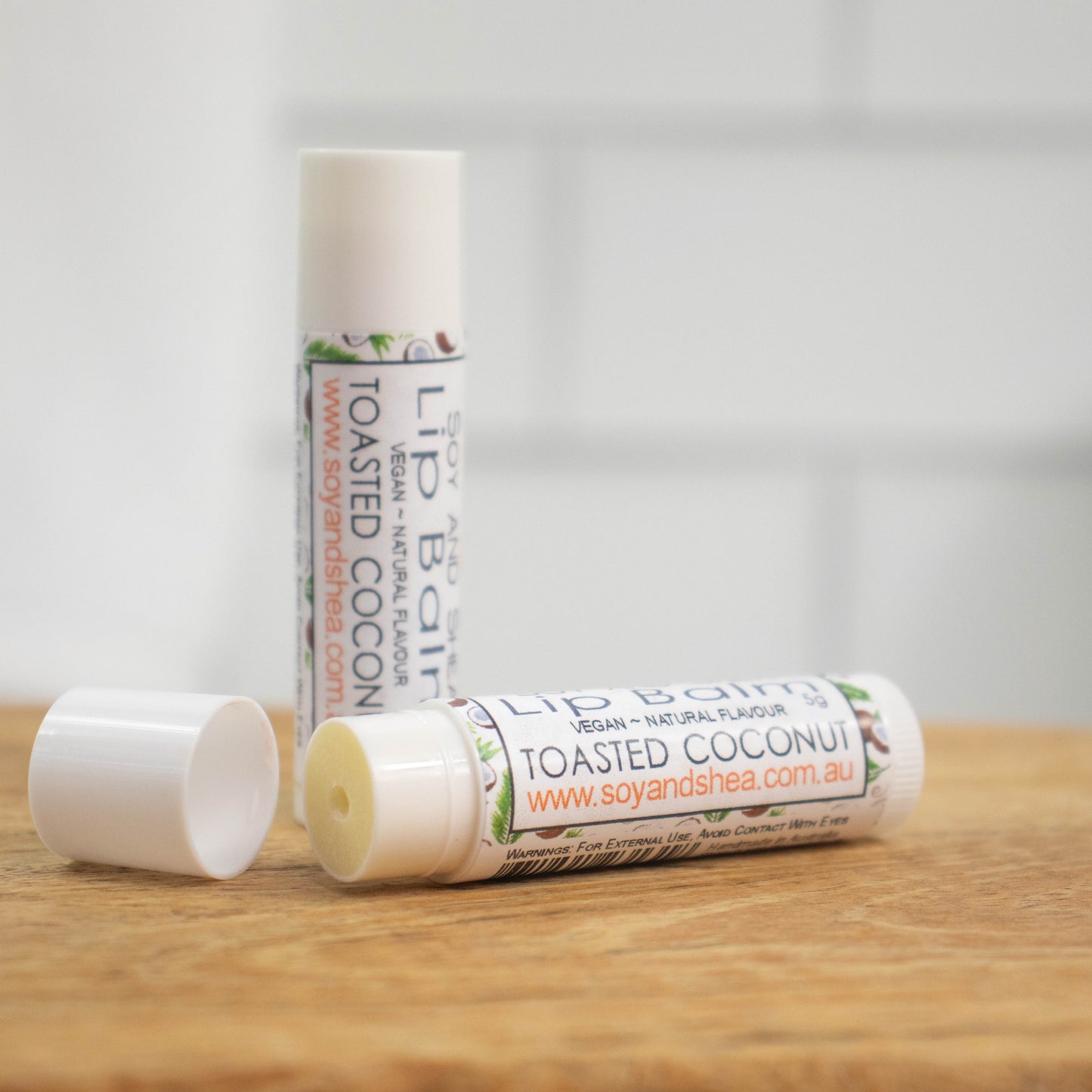 Two white tubes of lip balm are displayed on a wooden surface with blurred tiled background. One tube stands on its end while the other lays flat in front with the cap removed revealing an off-white balm. The label is white with a random pattern of coconut halves