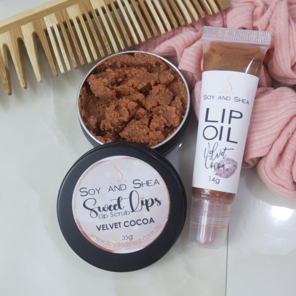 A small black tin with the lid removed revealing a chocolate brown Lip Scrub sits on a marble tile surface next to a small, clear tube of chocolate coloured Lip Oil. This rests againsts a wooden comb and pink scrunchie.
