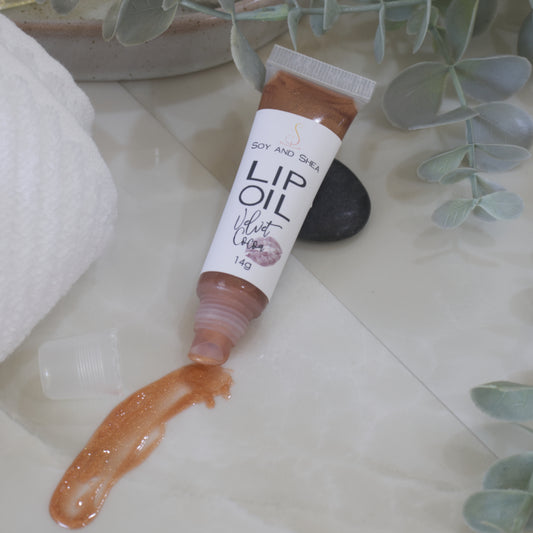 A tube of Lip Oil lies on a marble tile surface next to a white towel and some eucalyptus leaves. The tube is clear with a white product label and filled with a chocolate brown Lip oil. The cap is removed and a smear of Lip oil is on the bench showing the translucent effect it has