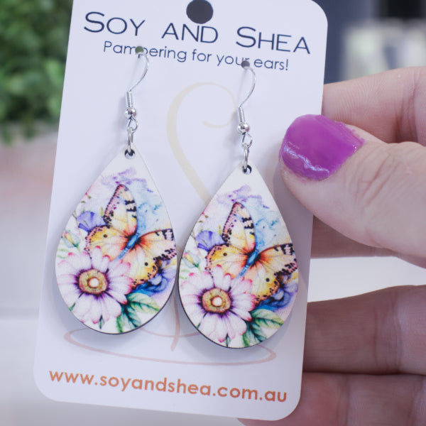 A hand holds a white card branded with SOY AND SHEA with a pair of earrings hanging on hooks. The earrings are teardrop shaped and feature a watercolour image of a large orange butterfly amound flower.