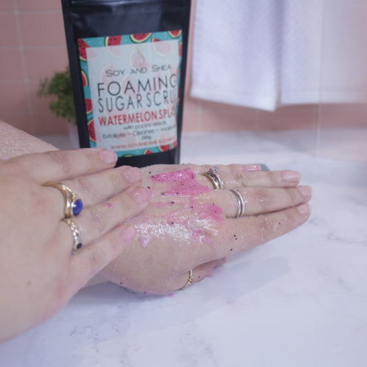 A hand applies the pink scrub with black poppy seeds to the back of the other hand. In the background sits a black pouch of the scrub with a label design of watermelon pieces against a tile background with white towel