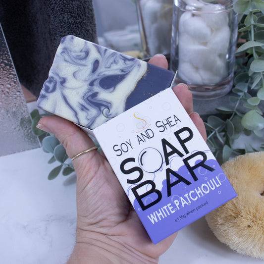A hand is holding onto a box that has a bar of soap coming out the top. The lower half of the box has a purple bubble shaped border, and the top is white with grey bubbles. The box shows it is soap and the scent. The soap is rectangular in shape with a layered rich purple bottom, with gold line and the top is white with purple swirls. In the background is a blurred bathroom counter top.