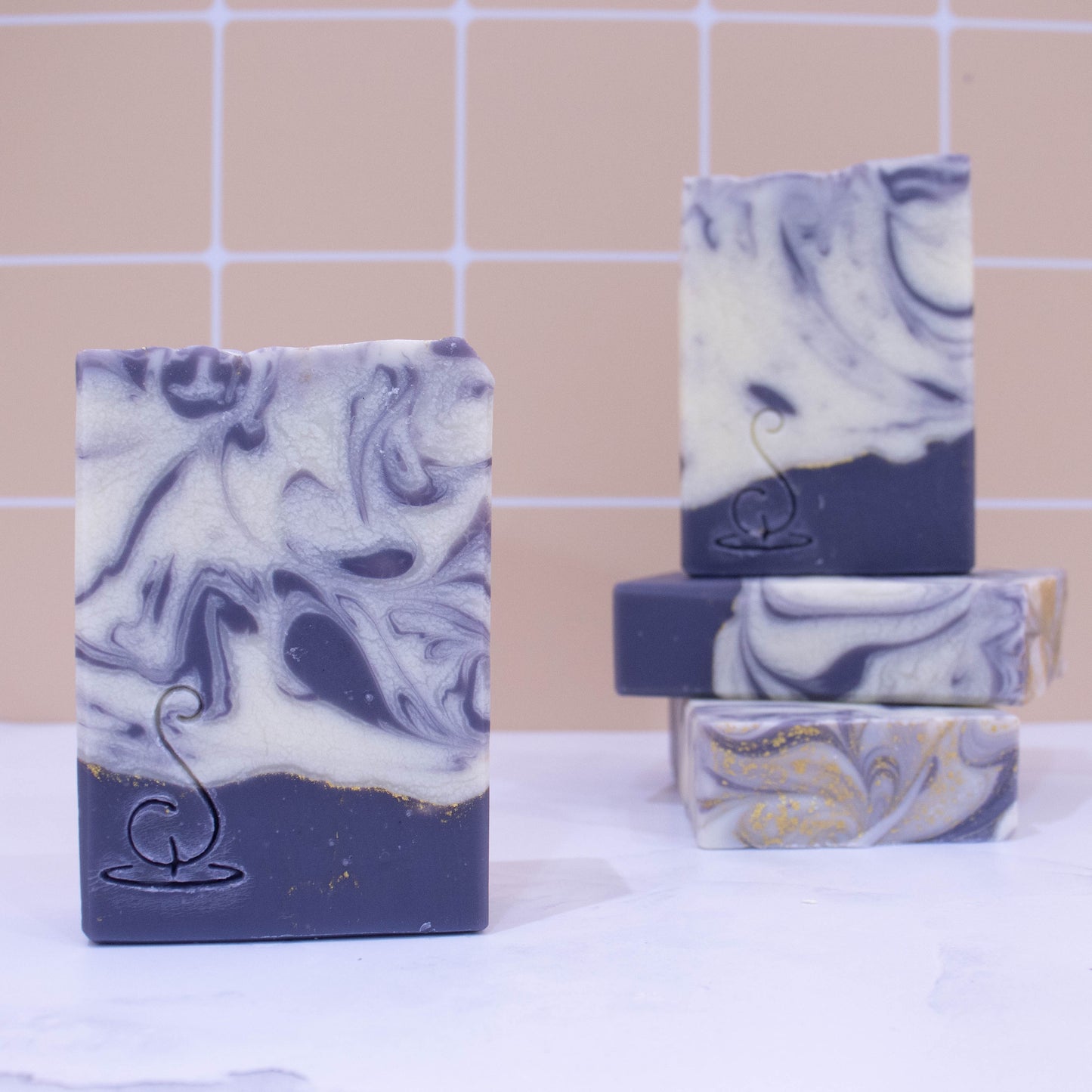 Four rectangular bars of soap sit on a marble bench each showing various design patterns from the same batch of soap. Two soaps lay flat but stacked upon each other showing a flat top. A third soap stands on these while a fourth is presented in front of the stack. The base of the soap is a rich purple with a thin gold line on top. This is followed by a white layer with rich purple swirls. All soaps have a similar pattern but all have slight differences
