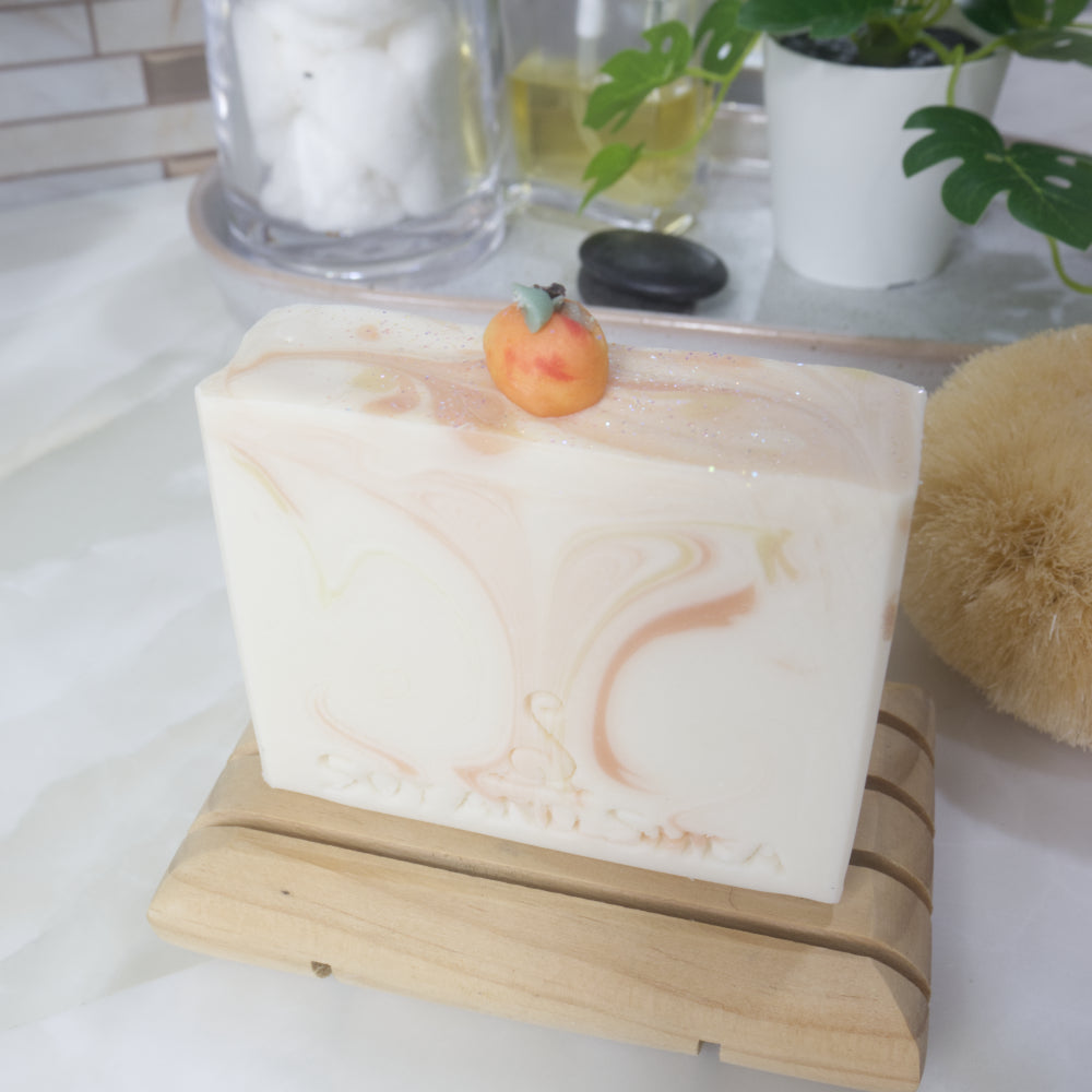 A soap stands on a wooden soap rack on a bathroom countertop. The soap is a cream colour with wisps of orange and yellow throughout. One top of the soap is a small replica of peach made with soap.