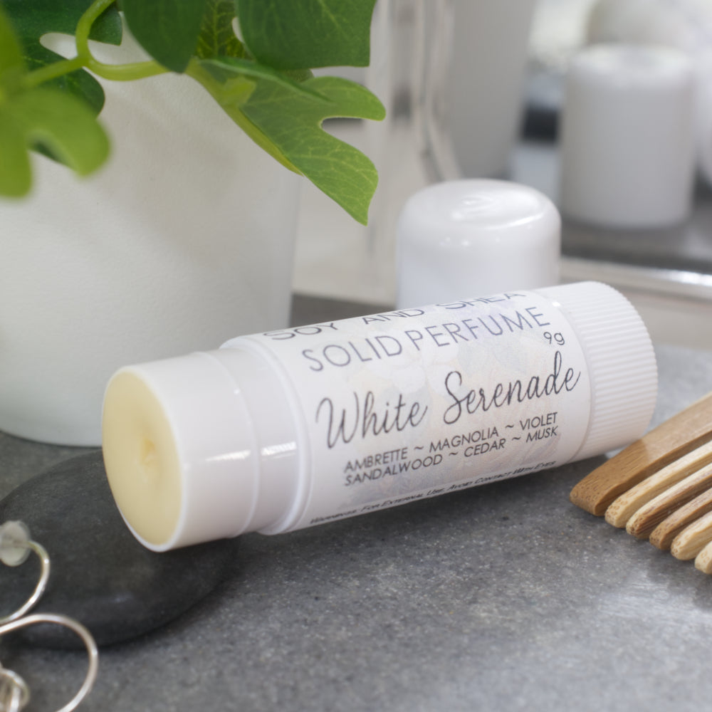 A white wind-up tube lays flat on a stone tray surrounded by accessories like earrings, watch and comb. The lid has been removed and placed behind the tube revealing a beige balm.