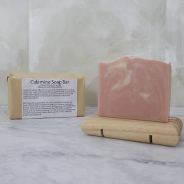 Pink soap bar on a wooden stand with a brown paper package labelled 'Calamine Soap Bar' on a marble background
