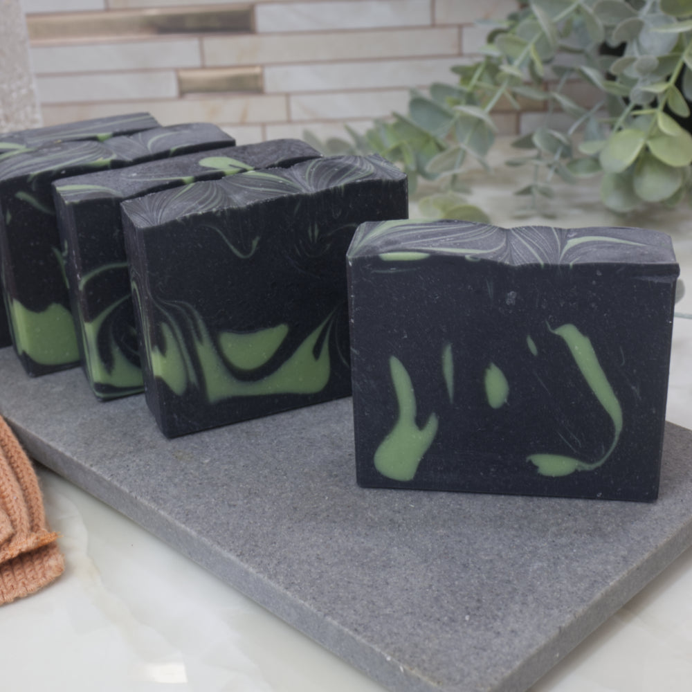 A selection of black and green marbled soap bars on a gray surface with a plant in the background. Each soap shows a different pattern of colours