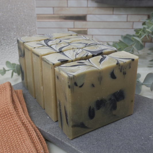 A row of soap bars on a gray stone surface surrounded by greenery and face cloth. The soaps are pale army green with black drop swirls