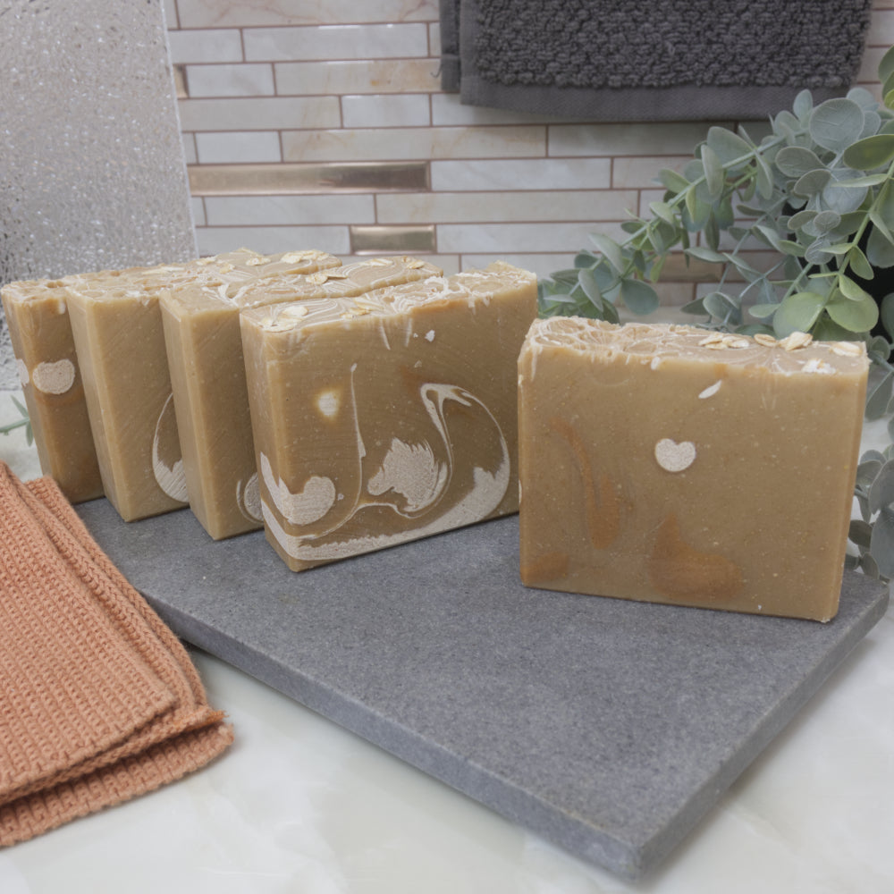 A selection of five soap bars sit on a stone tray surrounds by greenery and towels. The soaps sit one in front of the other and are slightly offset showing a unique design on each. The soaps are a light tan colour with swirls of white and gold.