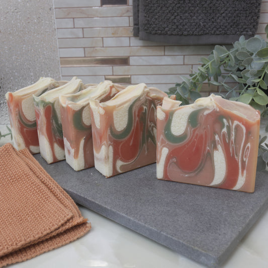 Marbled soap bars on a gray surface with a towel and plant in the background. The soaps sit one in front of the other, off set just slightly to see a peak of each design. The front soap shows a marbled design of orange, red, white and green