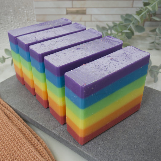 A row of rainbow-colored soap bars on a gray surface with a blurred background. The soaps feature six even layers of rainbow colour starting with red at the bottom and finishing with purple