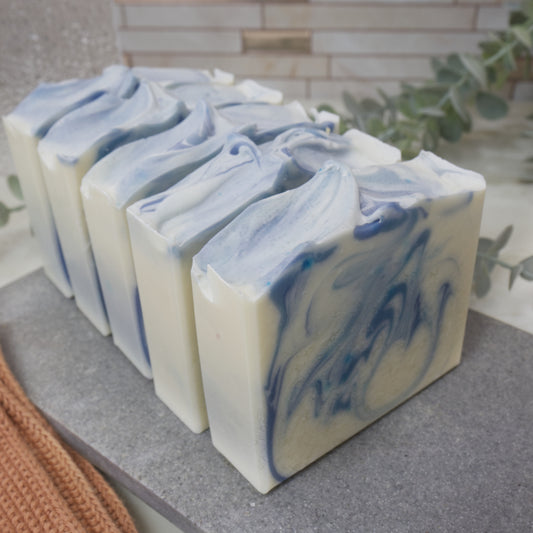 Five bars of soap sit one in front of the other with only the first soap showing the design. The soap has a main colour of white with a light and dark blue swirl. These all sit on stone tray on a bathroom counter top with greenery and towels
