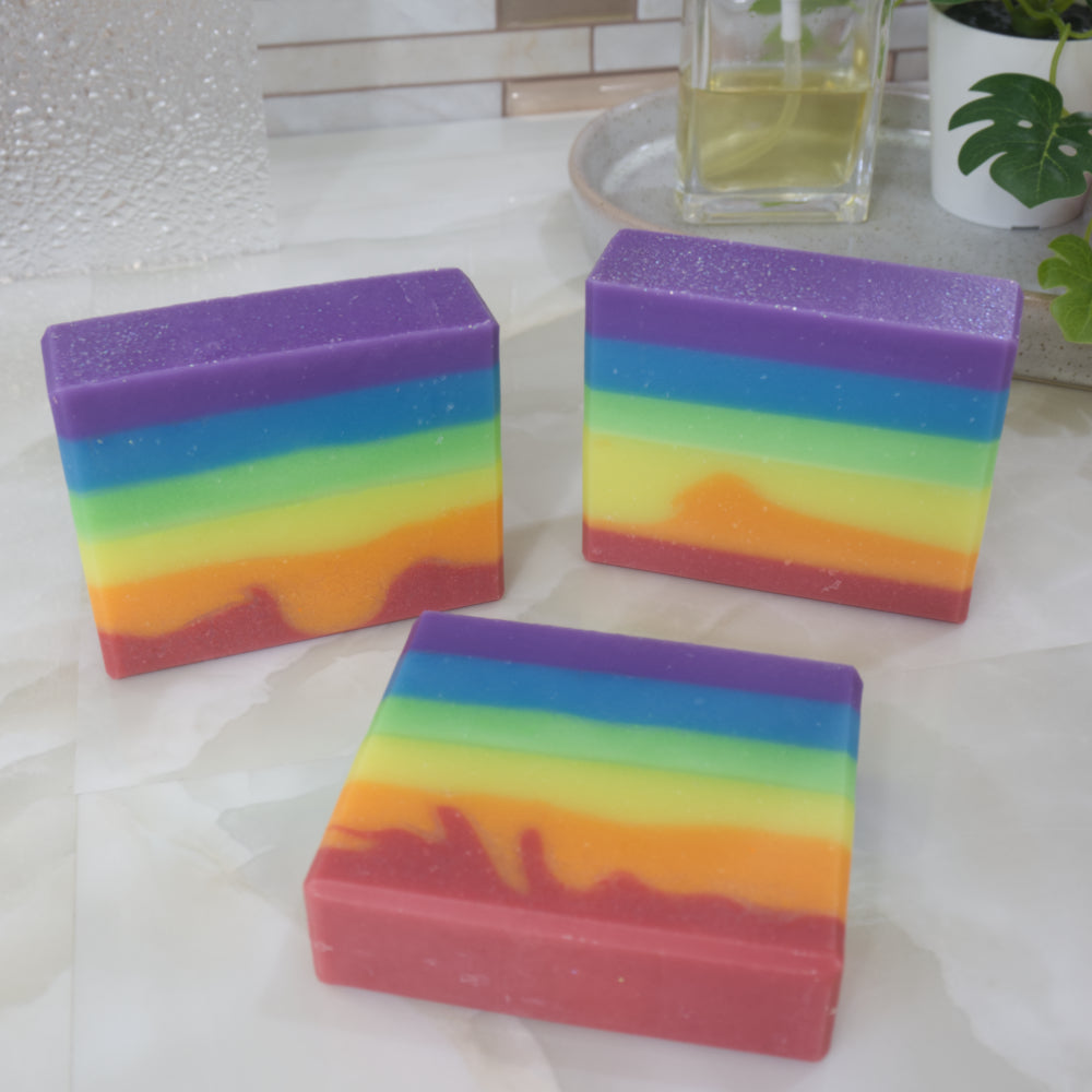 Three rainbow-colored soap bars on a white surface with a blurred background.