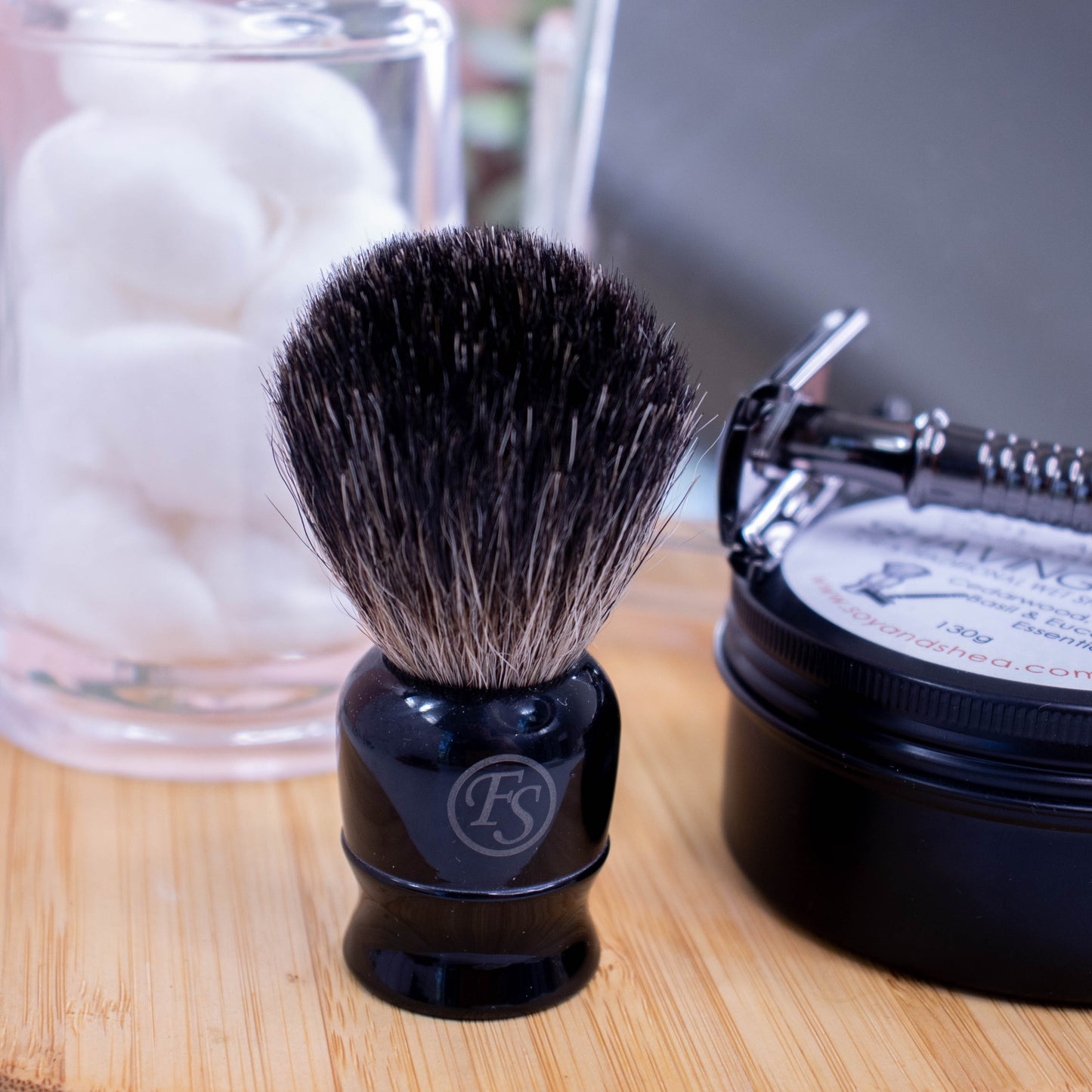Shaving brush stands on a wooden surface next to a jar of cotton balls and a tin of Shaving Soap with a razor resting on top. The brush as a squat black base with a logo of a circle with FS inside. The bristles are a long black colour with flecks of beige.