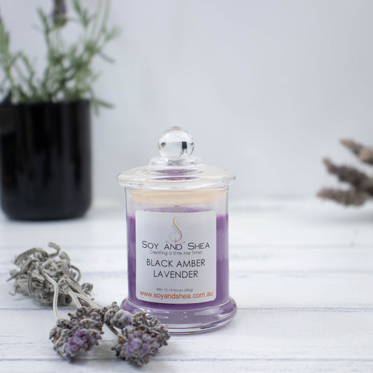 A small clear candle jar filled with purple wax sits at the front surrounded by lavender. The lid of the candle is dome shaped with a ball top. In the background is a pot of lavender growing