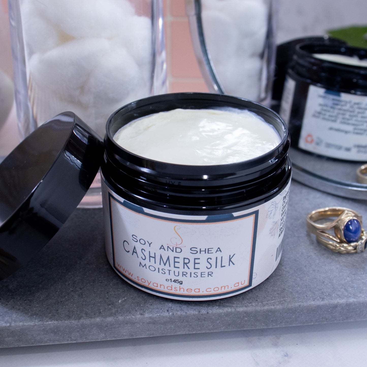 An open jar of moisturiser labelled CASHMERE SILK, sits on a grey stone surface next to jar of white cotton balls, some gold rings and a mirror reflecting the back of the jar. The jar is black and has a black lid resting on the side revealing an off-white cream inside.