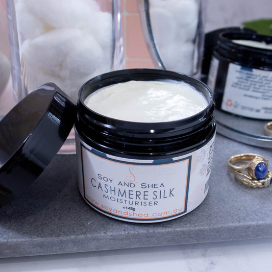 An open jar of moisturiser labelled CASHMERE SILK, sits on a grey stone surface next to jar of white cotton balls, some gold rings and a mirror reflecting the back of the jar. The jar is black and has a black lid resting on the side revealing an off-white cream inside.