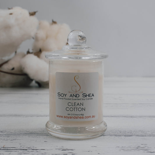 At the center of this photo sits a clear glass candle jar that features a lid with a small clear ball. It is filled with white wax and sits on a wooden bench with a bunch of cotton bolls in the background.