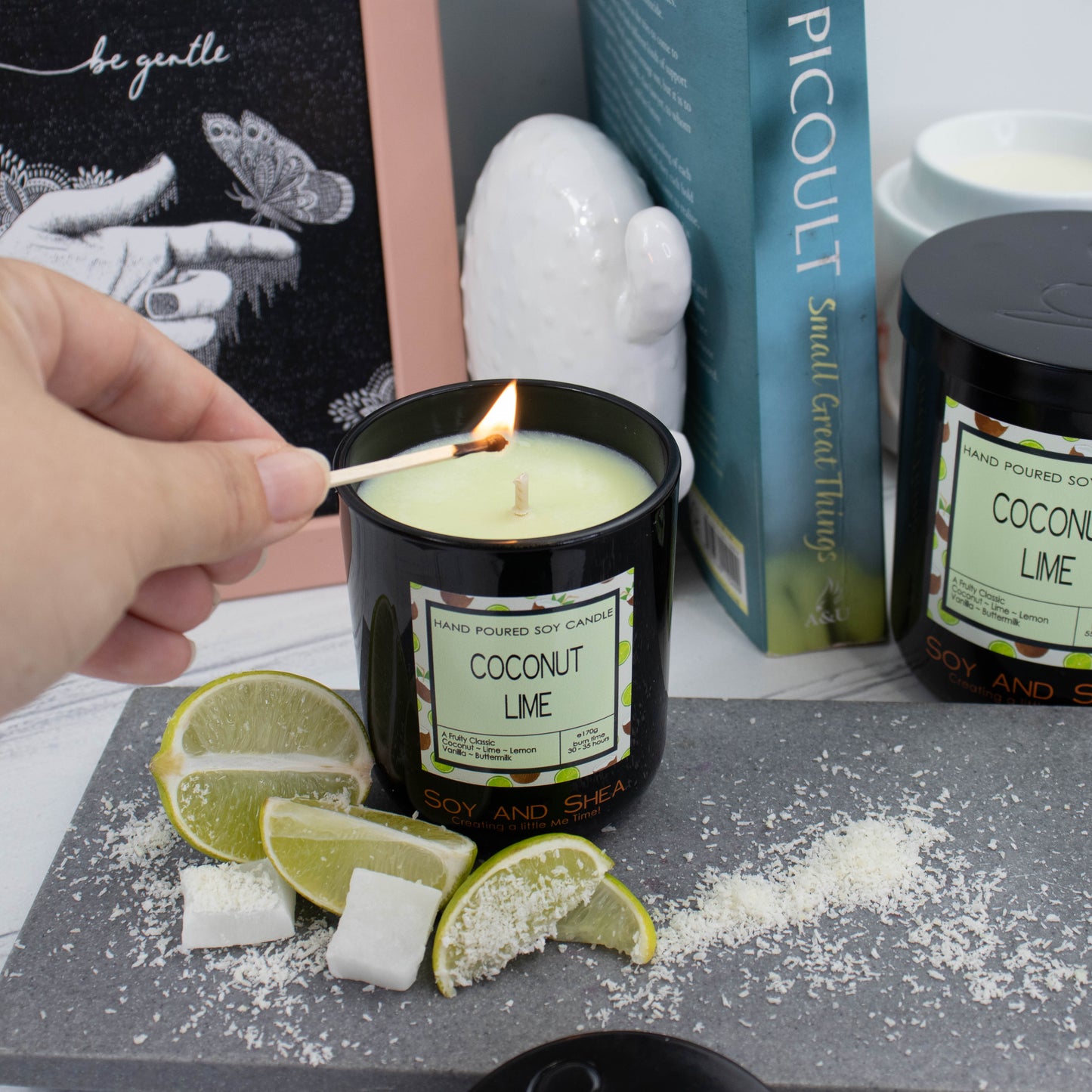A candle in a black glass jar filled with light green wax and a decorative product label is being lit with a match. Slices of lime and pieces of coconut surround the candle which sits on a stone tray. In the background is a picture frame, ornament and book along with a larger candle with a black metal lid.