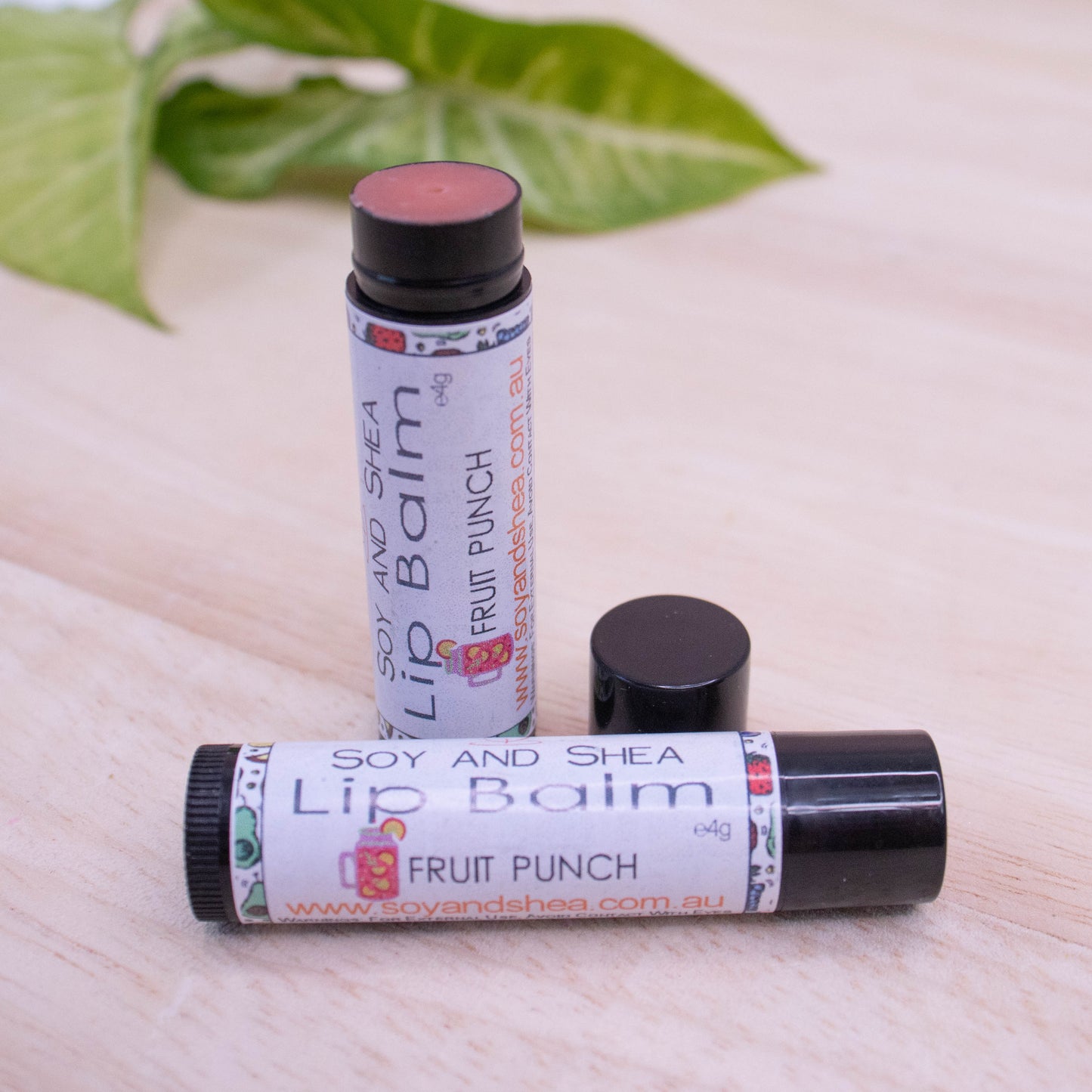 Two black tubes of lip balm displayed on a wooden surface with greenery blurred in the background. One tube lies flat across the surface displaying the flavour while the other tube stands upright behind it with the cap removed and placed to the side revealing a red coloured balm.
