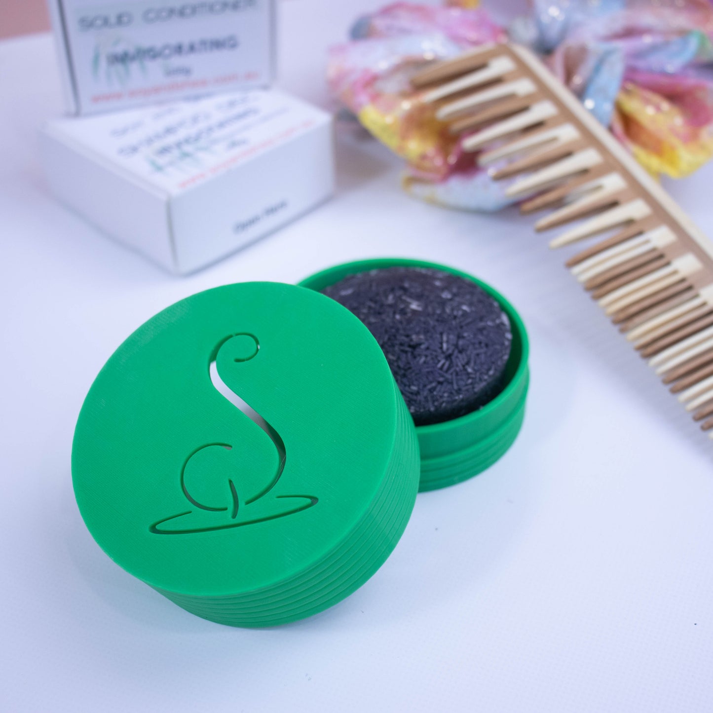 A Green cylindrical pot with the Soy and Shea logomark cut out in the top, sits on a white surface. The lid has been removed and is resting on the side of the base which holds a solid shampoo bar. In the background stands the shampoo packaging next to a wooden comb and colourful scrunchie with shiny gold dots.
