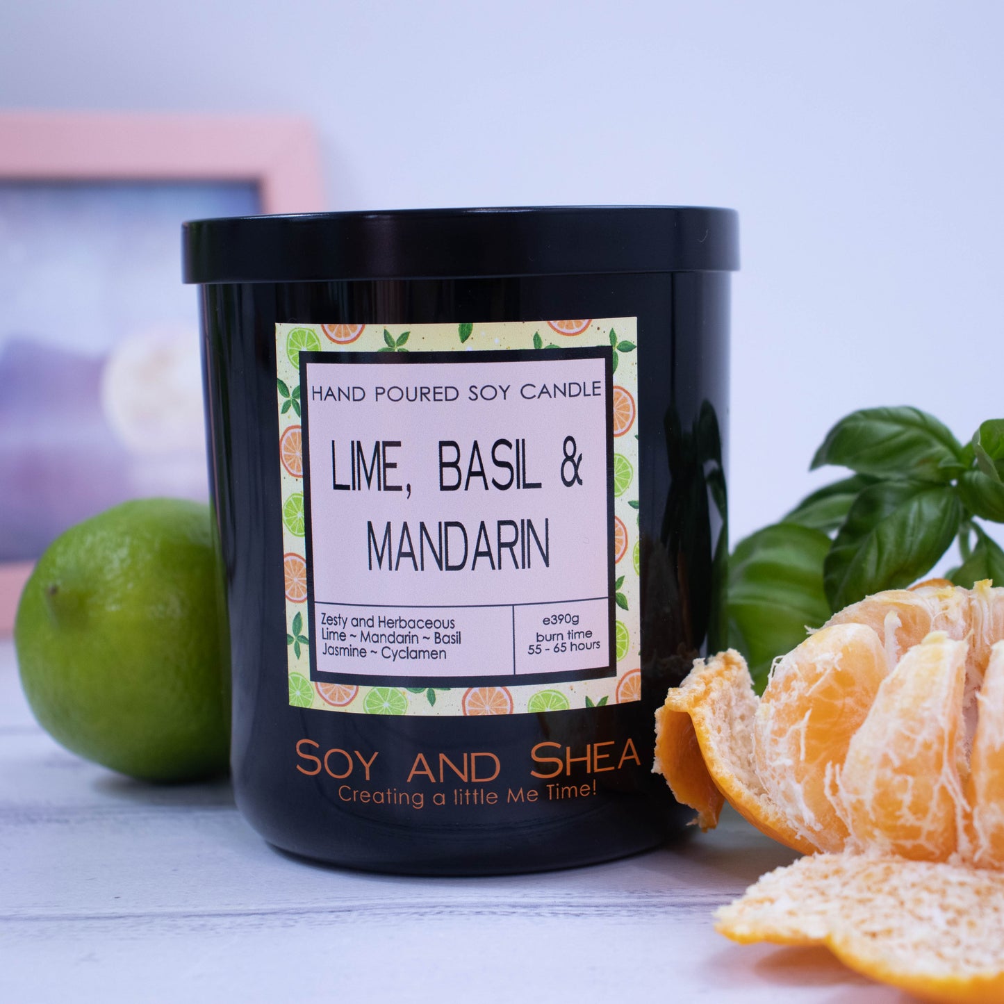 A large black glass jar with black metal lid sits on the wooden side. A decorative label is displayed on the front with information about the candle. The candle is surrounded by a peeled mandarin, whole lime and bunch of basil.