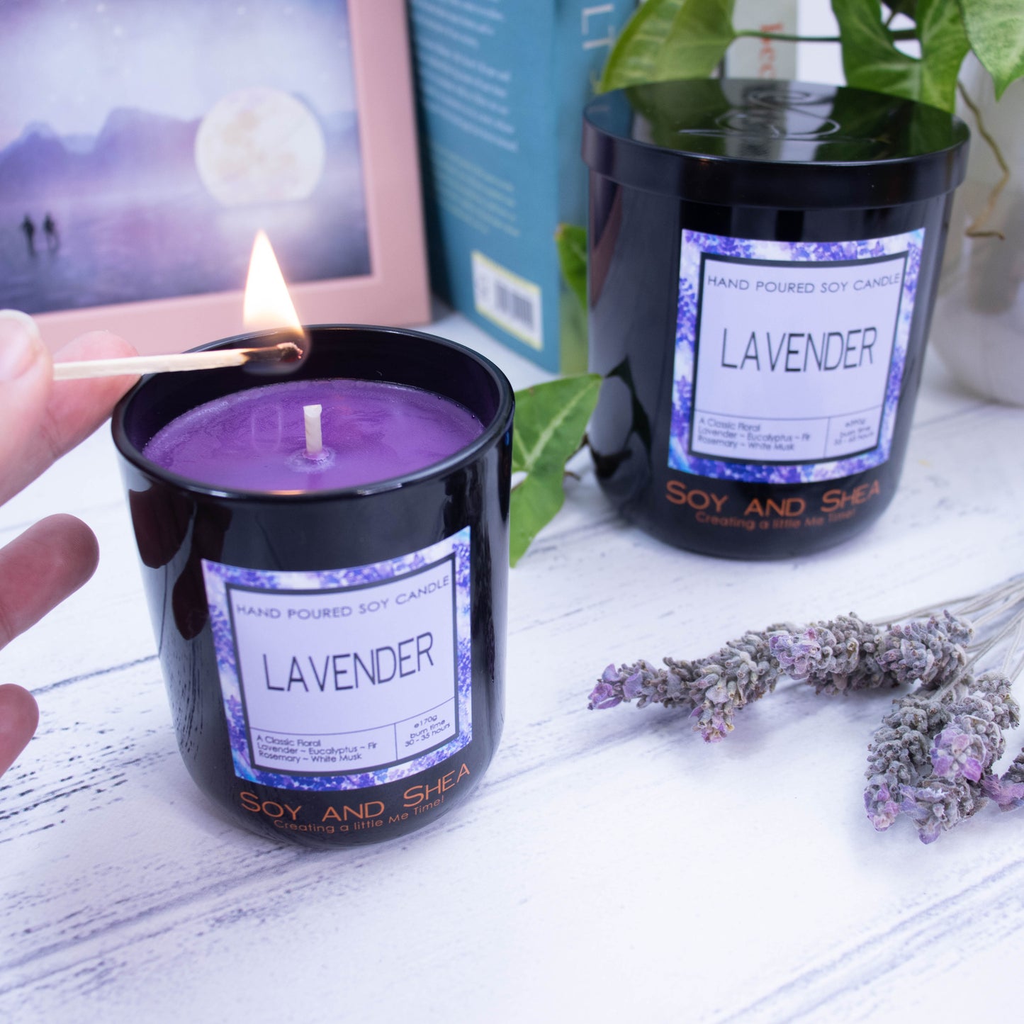 Two black glass candles sit on a wooden bench with one larger than the other. The larger candle is at the back and has the lid on and is surrounded by books, a picture and a plant. The smaller candle sits in front without a lid and is filled with purple wax and is about to be lit with a match
