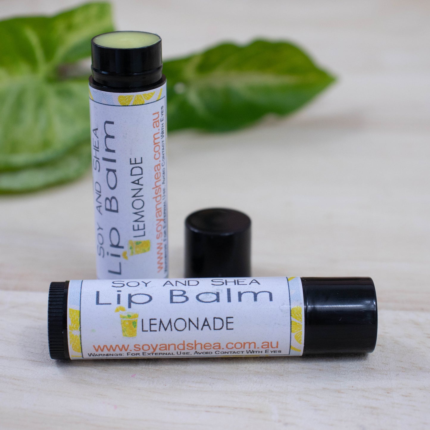 Two black tubes of lip balm displayed on a wooden surface with greenery blurred in the background. One tube lies flat across the surface displaying the flavour while the other tube stands upright behind it with the cap removed and placed to the side revealing a yellow balm.