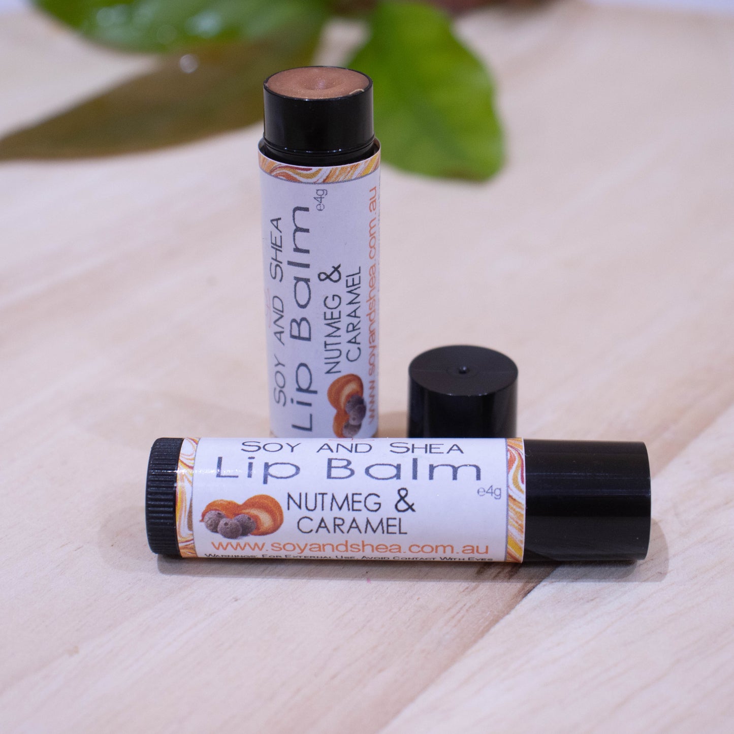 Two black tubes of lip balm displayed on a wooden surface with greenery blurred in the background. One tube lies flat across the surface displaying the flavour while the other tube stands upright behind it with the cap removed and placed to the side revealing a bronze coloured balm.
