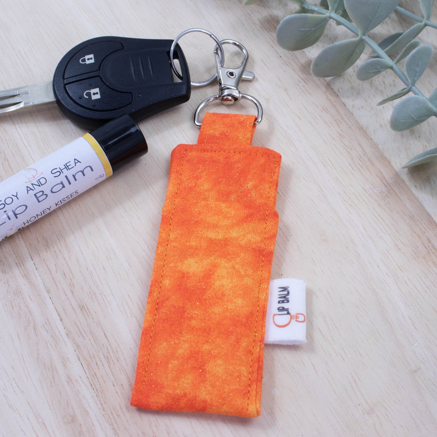 A clip balm holder lies on a wooden surface attached to a car key with a "lobster claw" clip. To the side is a tube of lip balm and some greenery. The Clip Balm has a bright orange mottled fabric with flecks of gold glitter.