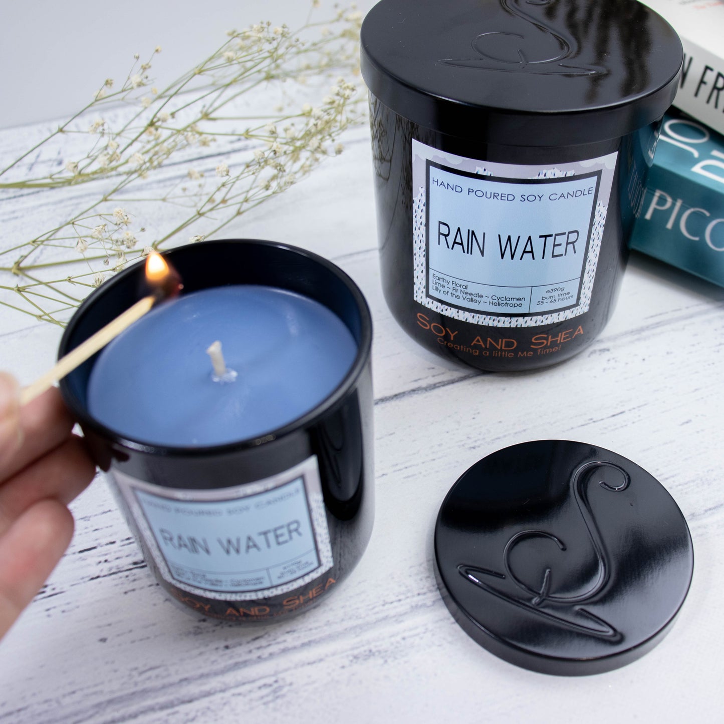 A large black jar candle with a decorative product label sits on a wooden surface next to stack of books and a sprig of babies breath. To the front, a small candle stands with its lid off and to the side revealing a dark blue wax. The lid is metal and has the logomark debossed omto the surface. A hand reaches in with a lit match to light the candle
