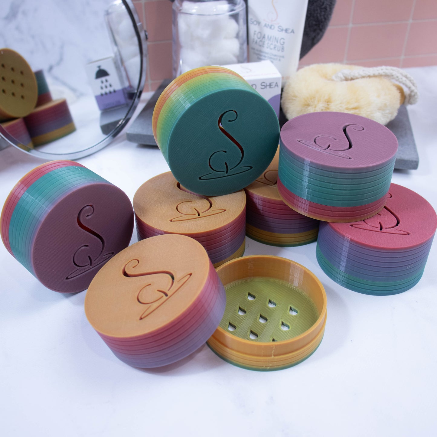 Seven Shampoo containers sit on a white bathroom countertop with mirror, body brush, cotton balls and tube of cream in the background. The containers are cylindrical with unique gradient colour patterns in muted purple, green, yellow and red. The Soy and Shea Logomark is cut out in the top of them. One container has the lid removed showing the base with waterdrop cutouts.